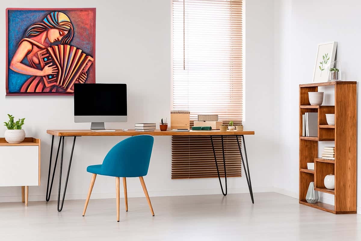 Oficina en casa moderna con un escritorio de madera, ese que parece invitarte a sentarte un rato largo. Encima, una computadora encendida y algunos cuadernos con notas dispersas, como si estuvieran esperando inspiración. La silla azul -cómoda y vibrante- destaca entre todo el mobiliario sobrio. En la pared cuelga 