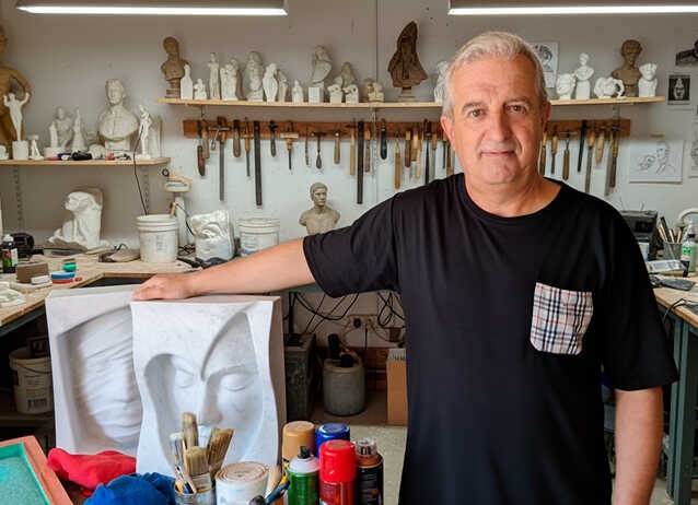 Antonio Gea Un hombre de cabello gris, con camiseta negra y bolsillo a cuadros, sonríe sutilmente junto a un bajo relieve blanco; atrás, estantes llenos de bustos curiosos, pinceles algo usados y tarros que parecen guardar secretos. Aquí podemos ver el Arte volumétrico contemporáneo de Antonio Gea