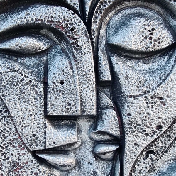 Primer plano de "Unidad Inquebrantable", esa escultura contemporánea que habla, casi susurra, sobre el amor eterno. Dos rostros humanos -imaginarios pero tan cercanos- se dibujan de perfil, con los ojos cerrados y los labios a un instante de encontrarse. La superficie no es lisa; más bien parece que cada marca y cada pequeño agujero guarda historias o silencios compartidos. Te dan ganas de pasar la mano y sentir ese relieve áspero, como si quisieras comprender todas las grietas del tiempo juntos. Porque sí: ahí, en esos detalles imperfectos, vive el verdadero significado del vínculo inquebrantable.